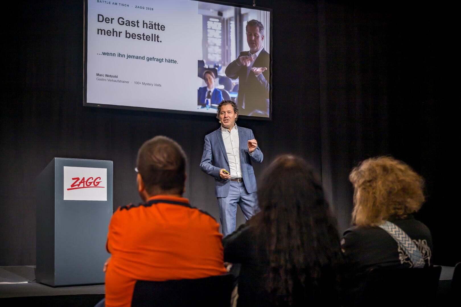 Ein Redner steht auf der Bühne vor einer Leinwand, auf der sein Foto und ein Text auf Deutsch zu sehen sind, während das Publikum zuhört.