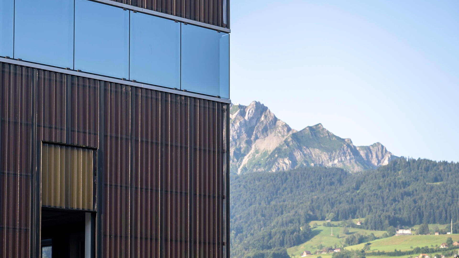 Auf der linken Seite steht ein modernes Gebäude mit reflektierendem Glas und vertikalen braunen Paneelen, im Hintergrund sind grüne Hügel und in der Ferne ein felsiger Berg unter einem klaren blauen Himmel zu sehen.