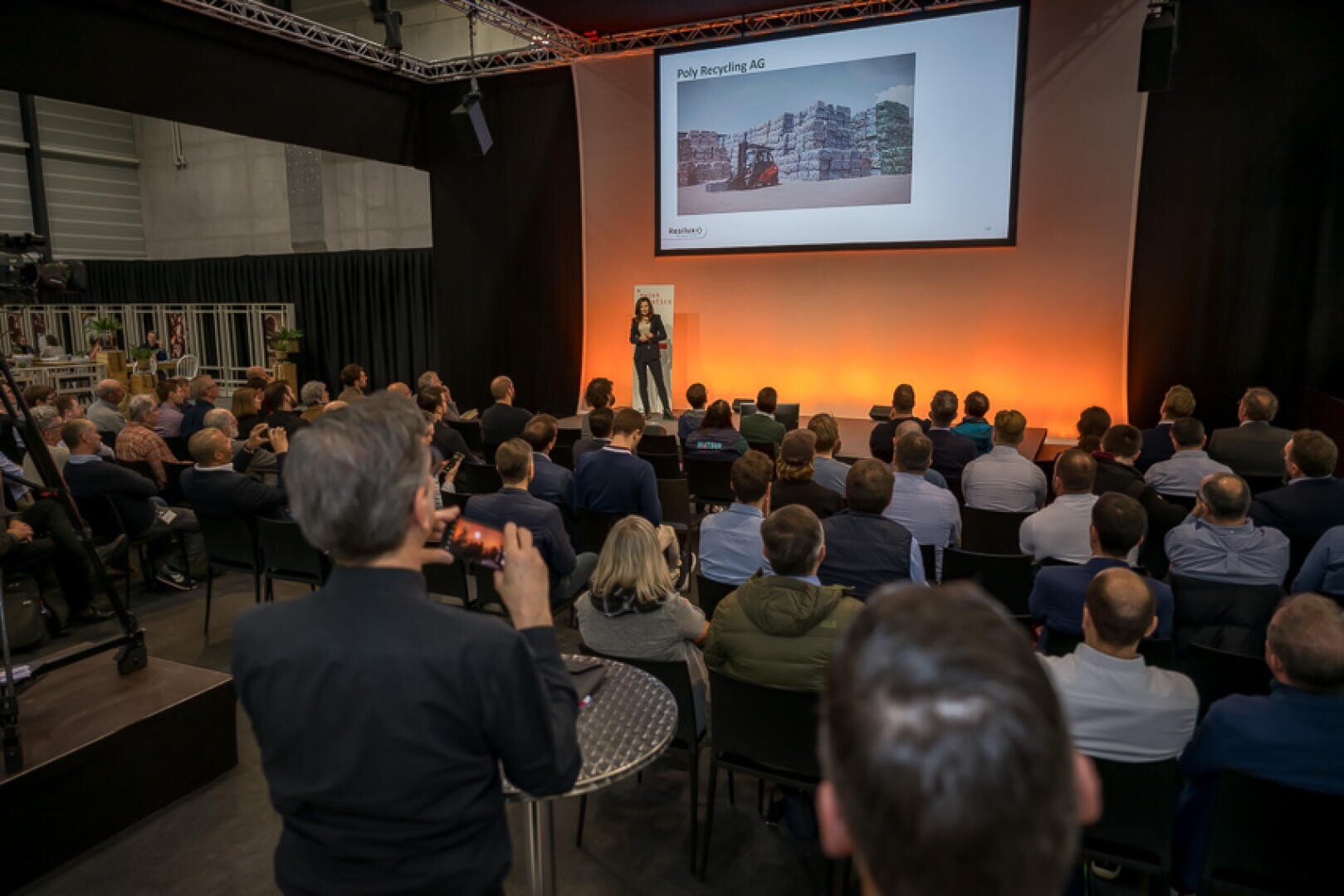 Eine Rednerin hält auf der Bühne einen Vortrag vor einem in Reihen sitzenden Publikum. Hinter ihr ist auf einer grossen Leinwand ein Bild mit gestapelten Recyclingmaterialien und der Aufschrift "Poly Recycling AG" zu sehen. Eine Teilnehmerin im Vordergrund nimmt die Veranstaltung auf.