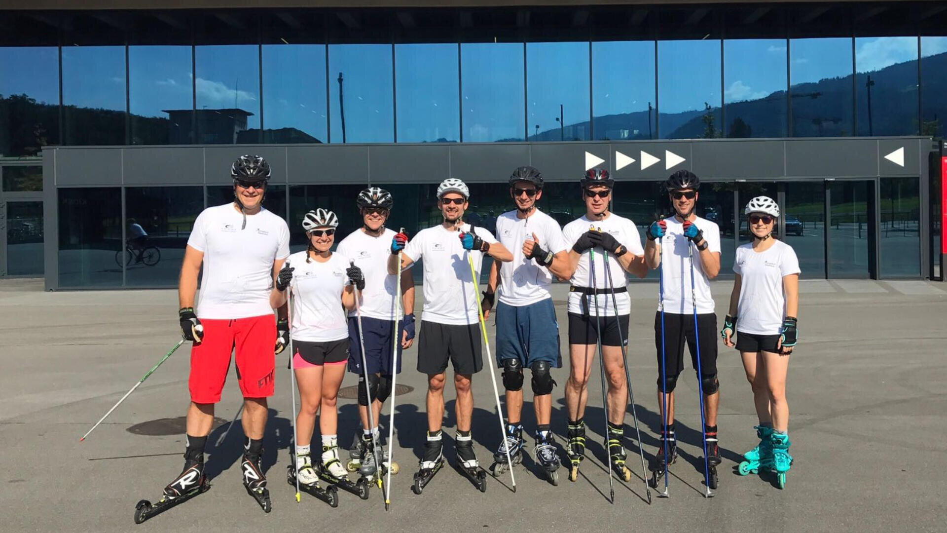 Eine Gruppe von sieben Personen mit Helmen, Sonnenbrillen und weißen Hemden steht in einer Reihe vor einem Gebäude und posiert an einem sonnigen Tag mit Rollskiern und Stöcken.