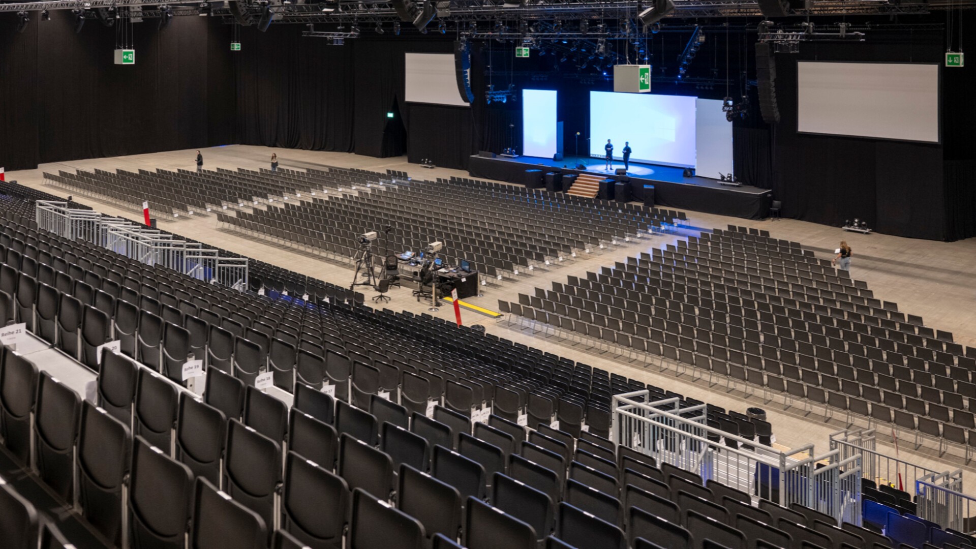 Ein großer, leerer Kongresssaal mit Reihen schwarzer Stühle vor einer Bühne. Die Bühne ist mit hellen Leinwänden, Beleuchtungsanlagen und einigen Leuten beim Aufbau ausgestattet. Industrielle Deckenbalken und Rohrleitungen sind darüber sichtbar.