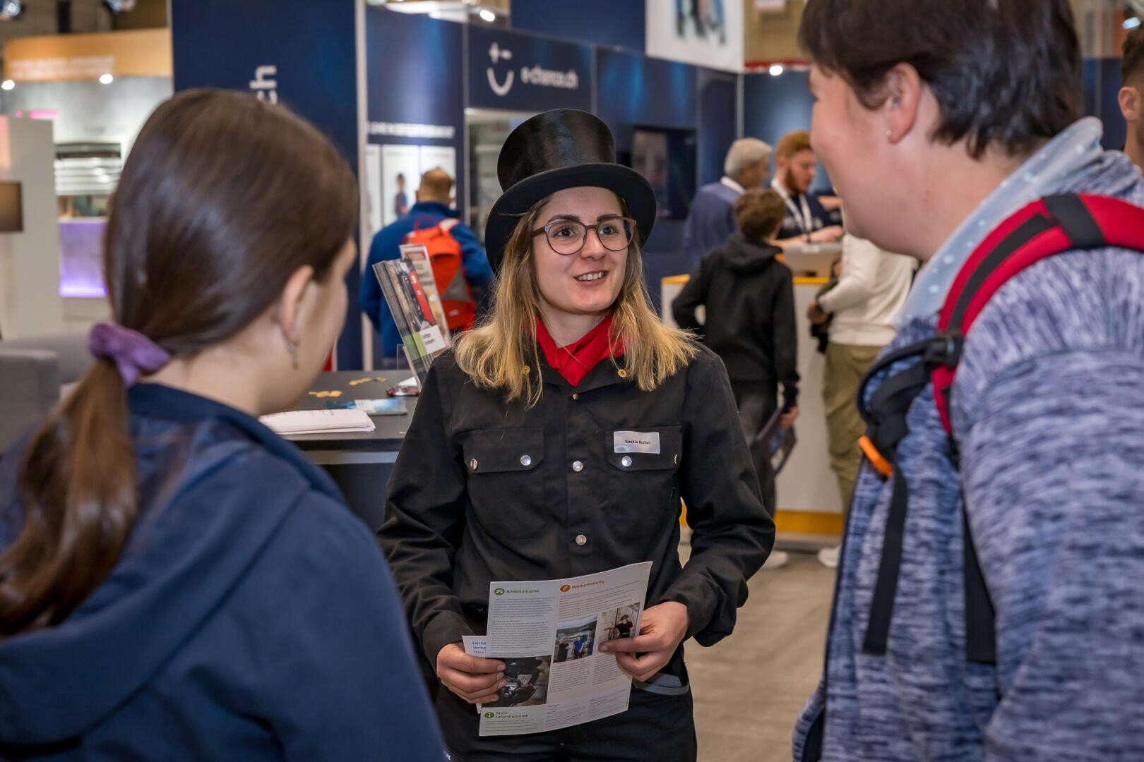 Eine Frau mit schwarzem Hut und Uniform unterhält sich bei einer Veranstaltung in einer Halle mit zwei Personen. Sie hält eine Broschüre in der Hand, und im Hintergrund sind andere Teilnehmer und Ausstellungsstände zu sehen.