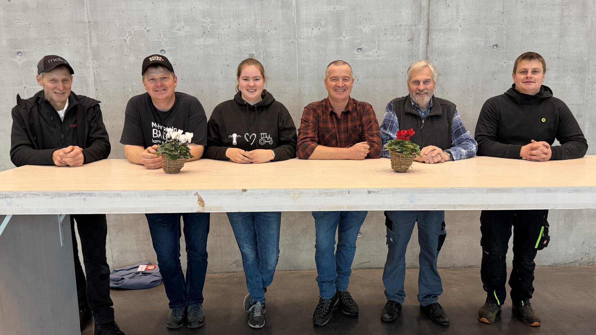 Sechs Personen sitzen nebeneinander an einem hellen Tresen mit kleinen Topfblumen und lächeln in die Kamera vor dem Hintergrund einer schlichten Betonwand.