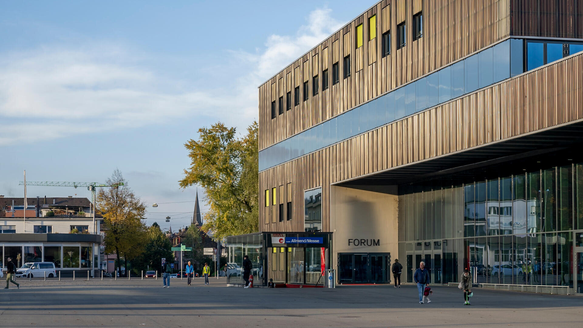 Modernes Gebäude mit der Aufschrift FORUM, großen Glasfenstern und Holzvertäfelung, neben einem offenen Platz gelegen. In der Nähe gehen Menschen spazieren und radeln. Im Hintergrund sind Bäume und Stadtgebäude unter einem teilweise bewölkten Himmel zu sehen.