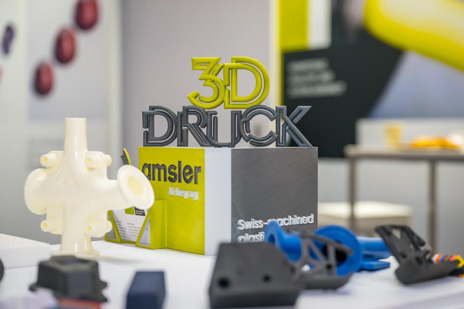 Ein Display mit einem 3D-gedruckten Schild mit der Aufschrift 3D DRUCK inmitten verschiedener 3D-gedruckter Objekte in verschiedenen Formen und Farben, das auf einem Tisch auf einer Ausstellung oder Messe aufgestellt ist.