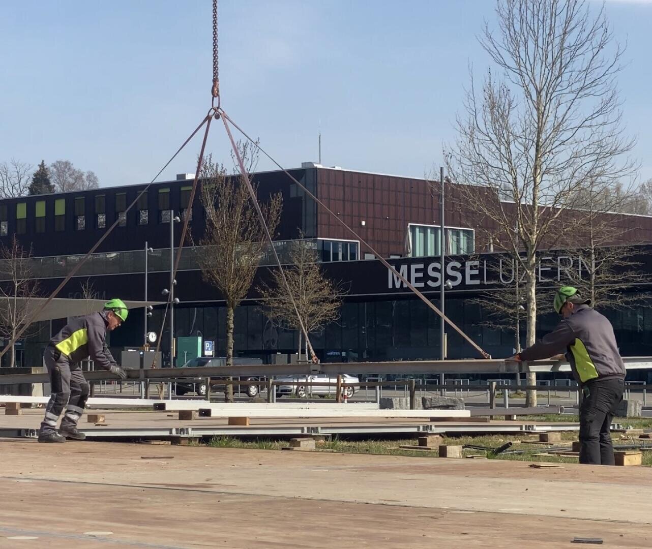 Zwei Bauarbeiter mit Schutzausrüstung führen einen großen Metallträger, der von einem Kran vor einem modernen Gebäude mit der Aufschrift "MESSE LUZERN" angehoben wird. Im Hintergrund sind Bäume und ein klarer Himmel zu sehen.