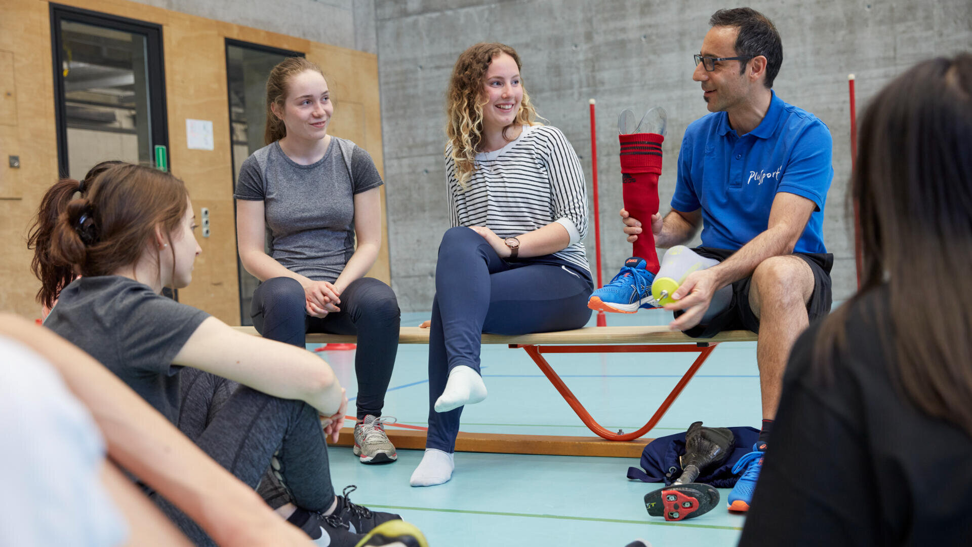 Eine Gruppe junger Leute sitzt auf einer Turnbank und dem Boden, hört lächelnd zu, während ein blau gekleideter Trainer mit Tennisschuh und Ball etwas erklärt. Die Szene spielt in einer modernen Sporthalle.