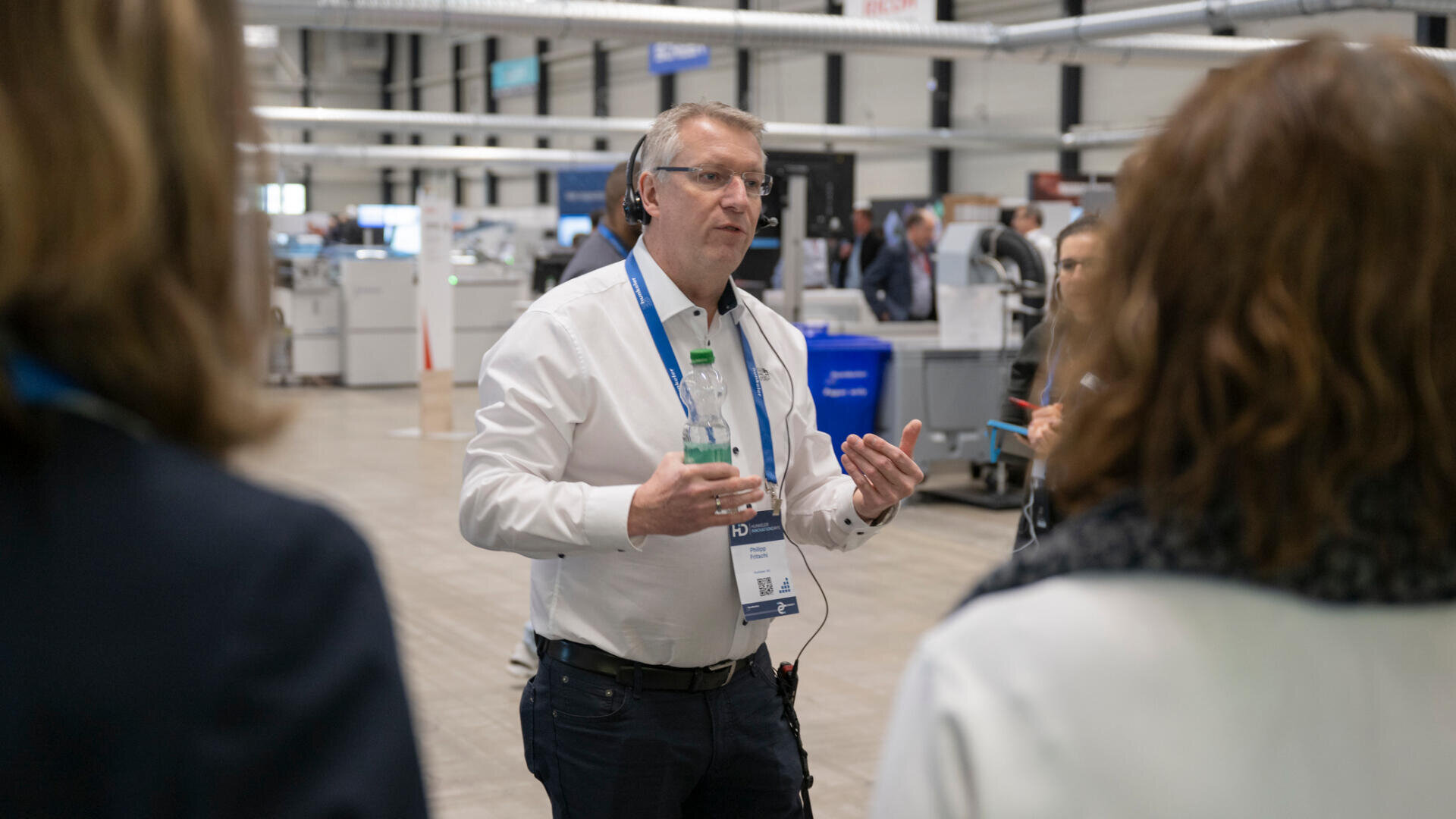 Ein Mann mit Brille und Konferenzausweis spricht in einem Industrie- oder Konferenzumfeld zu einer kleinen Gruppe von Menschen, hält eine Wasserflasche in der Hand und gestikuliert mit den Händen.