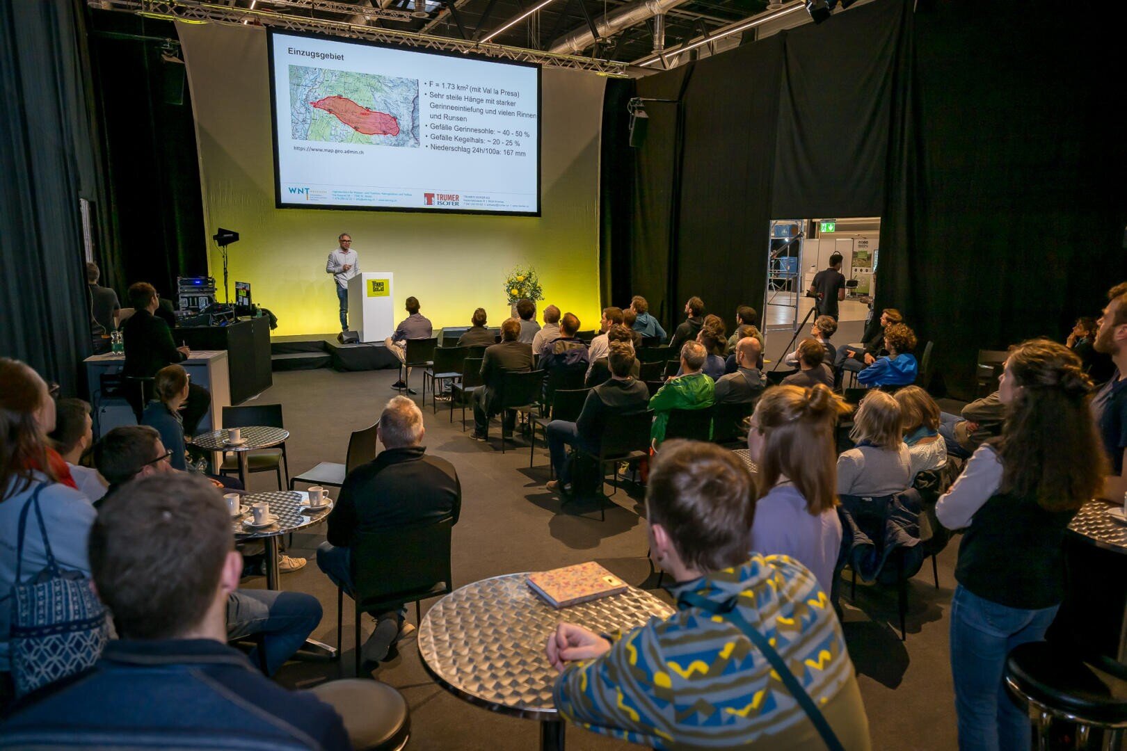 Ein Redner präsentiert dem Publikum, das auf Stühlen und Hockern in einem schwach beleuchteten Veranstaltungsraum mit schwarzen Vorhängen und runden Tischen sitzt, eine Folie mit einer Karte und einem Text.