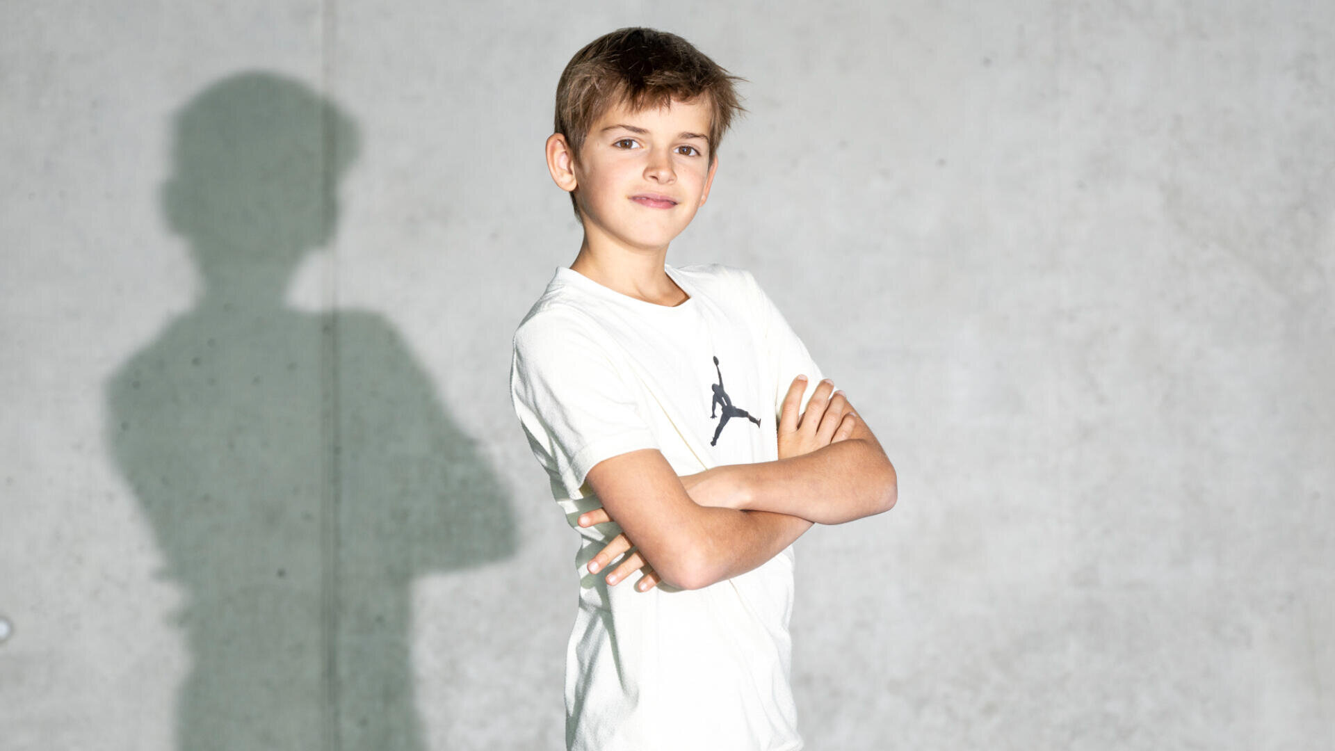Ein kleiner Junge mit kurzen braunen Haaren steht selbstbewusst mit verschränkten Armen da und trägt ein weißes T-Shirt mit einem kleinen schwarzen Logo. Er blickt in die Kamera und lächelt, wodurch ein deutlicher Schatten auf die schlichte graue Wand hinter ihm fällt.