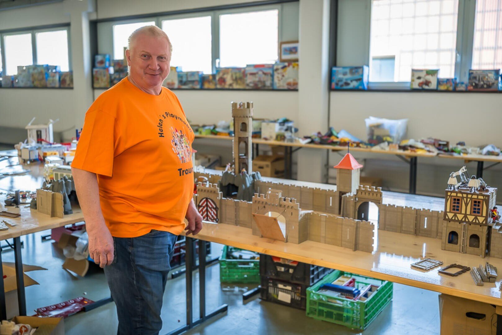Ein lächelnder Mann in einem orangefarbenen T-Shirt steht neben einer großen, detaillierten Modellburg auf einem Tisch in einem hellen Raum, der mit Kisten, Spielzeug und Baumaterialien gefüllt ist.