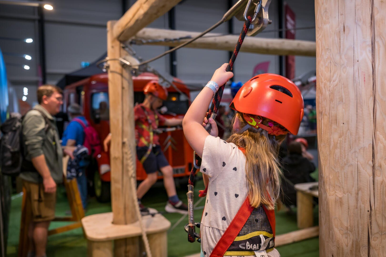 Ein Kind, das einen Helm und einen Klettergurt trägt, bereitet sich auf das Klettern in einem Indoor-Seilgarten vor, während andere Personen im Hintergrund zusehen und mitmachen. Der Schauplatz scheint ein Abenteuerzentrum oder eine Indoor-Veranstaltung zu sein.