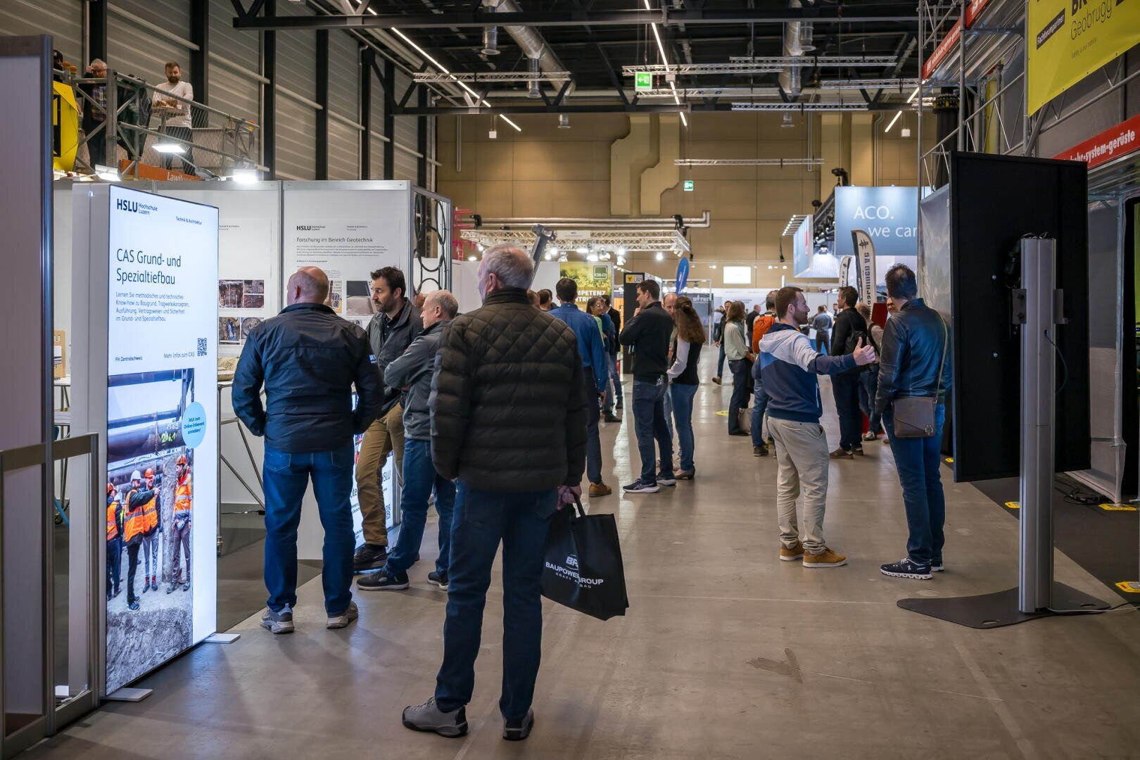 Auf einer Indoor-Messe oder -Ausstellung mit verschiedenen Ständen, Informationsdisplays und Bannern gehen Menschen umher und interagieren. Die Umgebung wirkt geschäftig und professionell, mit heller Beleuchtung und einem modernen, industriellen Interieur.