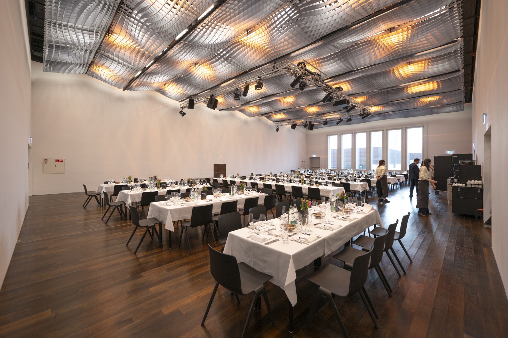 Ein geräumiger, moderner Bankettsaal mit langen, in Reihen angeordneten Tischen ist für ein formelles Abendessen gedeckt. Der Raum hat Holzböden, große Fenster und eine geometrische Decke mit dekorativer Beleuchtung. Mehrere Personen bereiten den Raum vor.