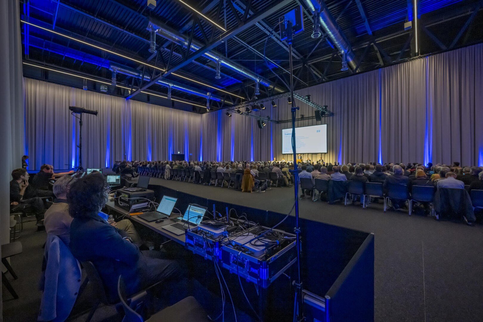 Ein großes Publikum nimmt an einer Konferenz in einem geräumigen, modernen Saal mit blauer Beleuchtung teil. Im Vordergrund sind technische Geräte und Bediener zu sehen, während ein Redner auf der Bühne auf einen Bildschirm spricht.