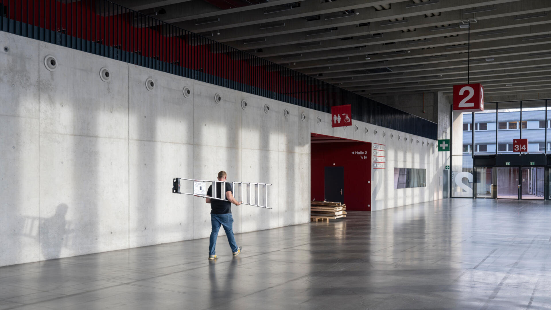Eine Person trägt eine lange Leiter durch einen geräumigen, modernen Flur mit Betonwänden, großen Fenstern und Wegweisern mit der Zahl 2. Sonnenlicht wirft Schatten auf Boden und Wände.