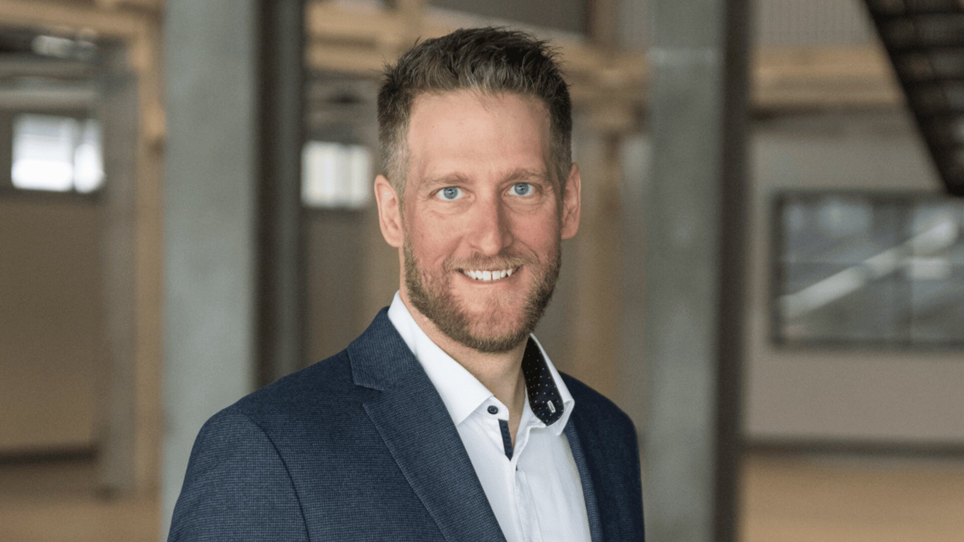 Ein Mann mit kurzen Haaren und Bart, der einen blauen Blazer und ein weißes Hemd trägt, lächelt in die Kamera in einer modernen Inneneinrichtung mit Betonsäulen und großen Fenstern im Hintergrund.
