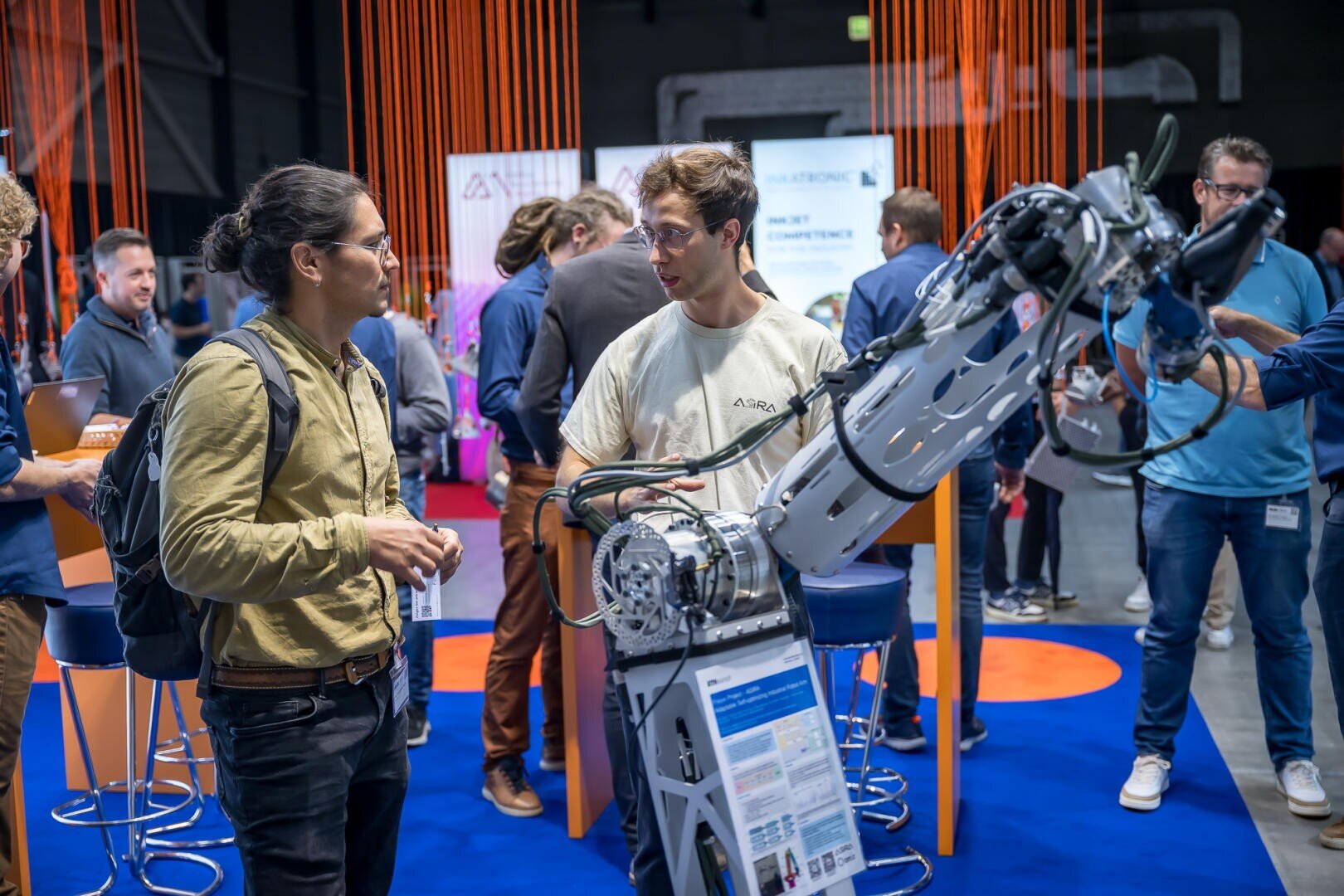 Zwei Männer unterhalten sich neben einem großen Roboterarm auf einer belebten Technologiemesse. Im Hintergrund mischen sich Menschen zwischen orangefarbenen Kordeldekorationen, Informationsständen und blauen Matten. Ein Mann hat einen Rucksack in der Hand, der andere trägt eine Brille und ein weißes Hemd.