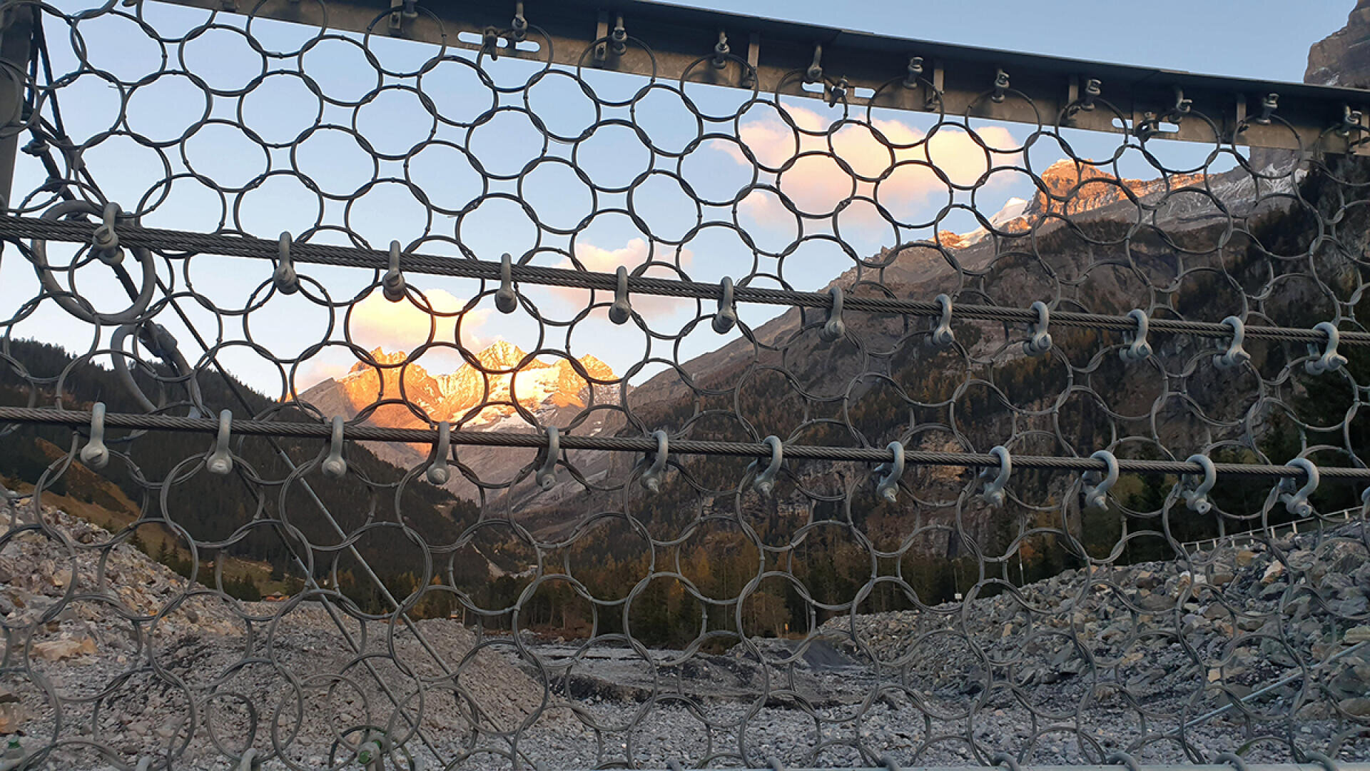 Im Vordergrund ein Maschendrahtzaun mit felsigen Bergen und Bäumen im Hintergrund, beleuchtet vom warmen Licht der untergehenden oder aufgehenden Sonne.