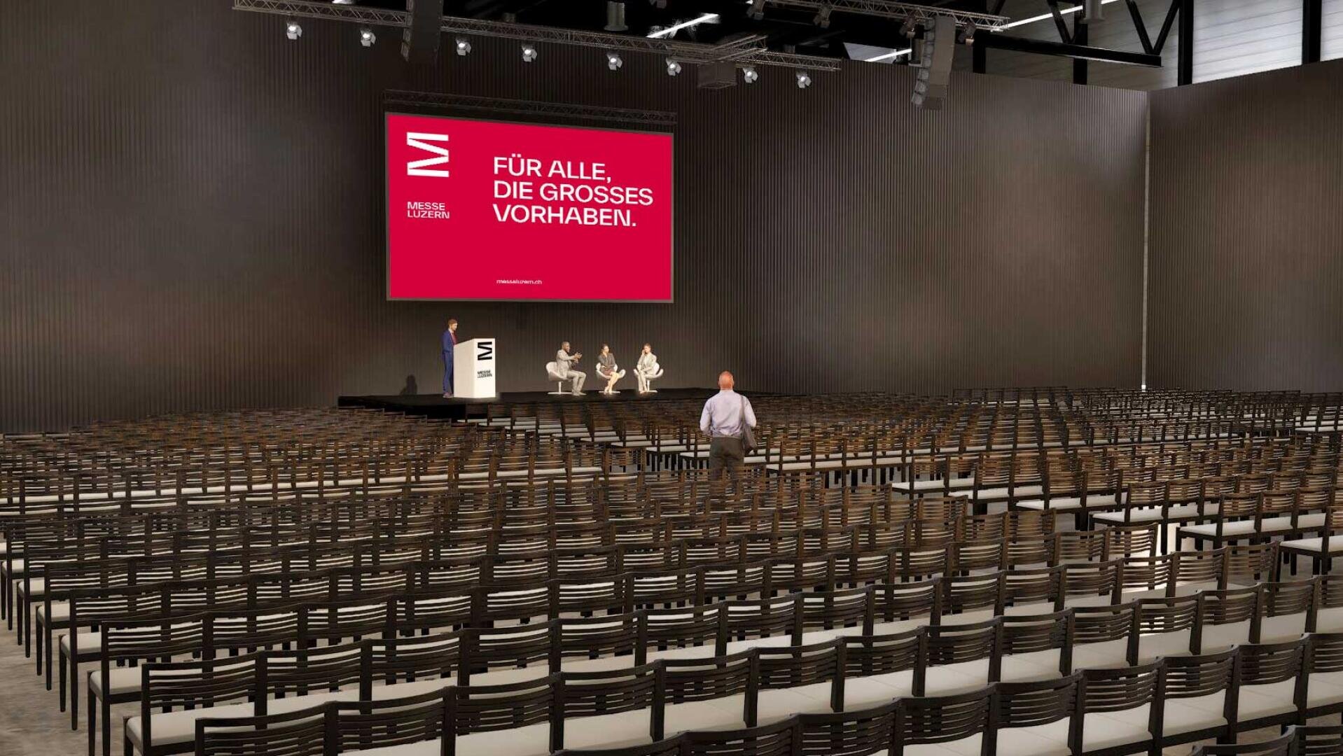 Ein größtenteils leerer Konferenzsaal mit Stuhlreihen, einer Bühne mit vier Sitzplätzen und einem großen roten Bildschirm, auf dem ein deutscher Text angezeigt wird: Für alle, die Großes vorhaben. Eine Person steht im Zuschauerbereich.