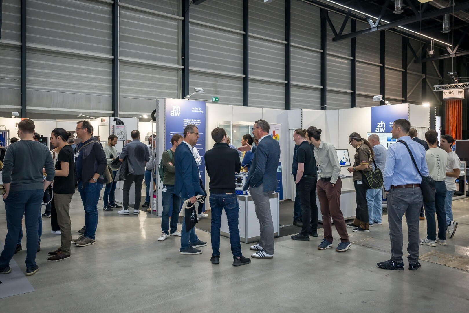 Eine Gruppe von Menschen steht und unterhält sich auf einer Ausstellung oder Messe in einer Halle, mit Ausstellungsständen im Hintergrund und verschiedenen Informationsmaterialien auf Tischen. Die Atmosphäre ist professionell und ungezwungen.