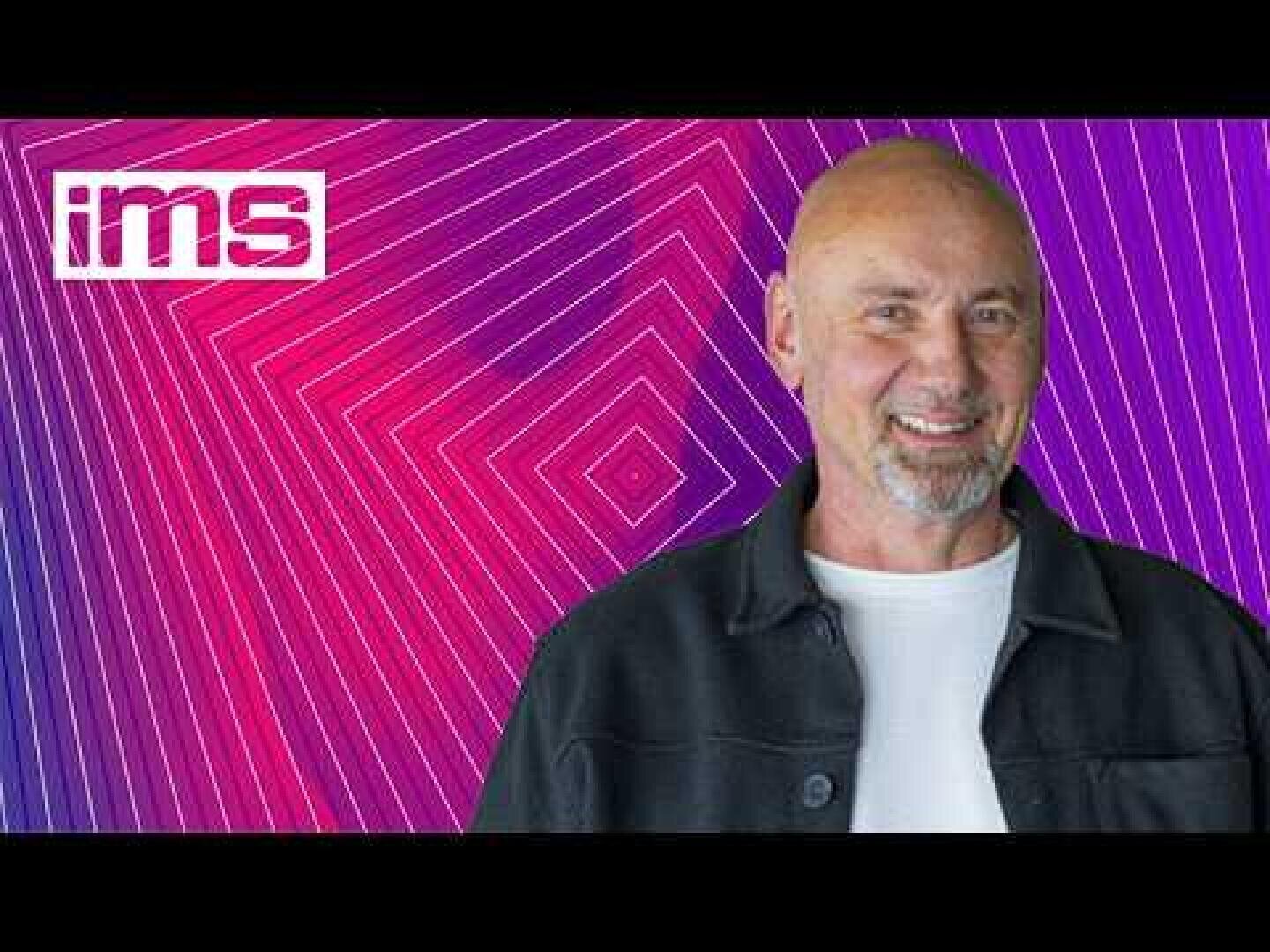 Ein lächelnder, kahlköpfiger Mann in einem weißen T-Shirt und einer schwarzen Jacke, der vor einem violetten und rosa geometrischen Hintergrund steht, in dessen Ecke ein IMS-Logo zu sehen ist.