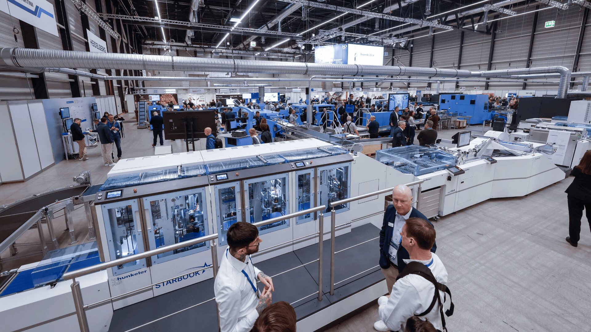 Eine geräumige, moderne Industrieanlage mit großen blau-weißen Maschinen und Menschengruppen. Manche unterhalten sich, andere beobachten Geräte oder Vorführungen. Die Szene ist gut beleuchtet und belebt.