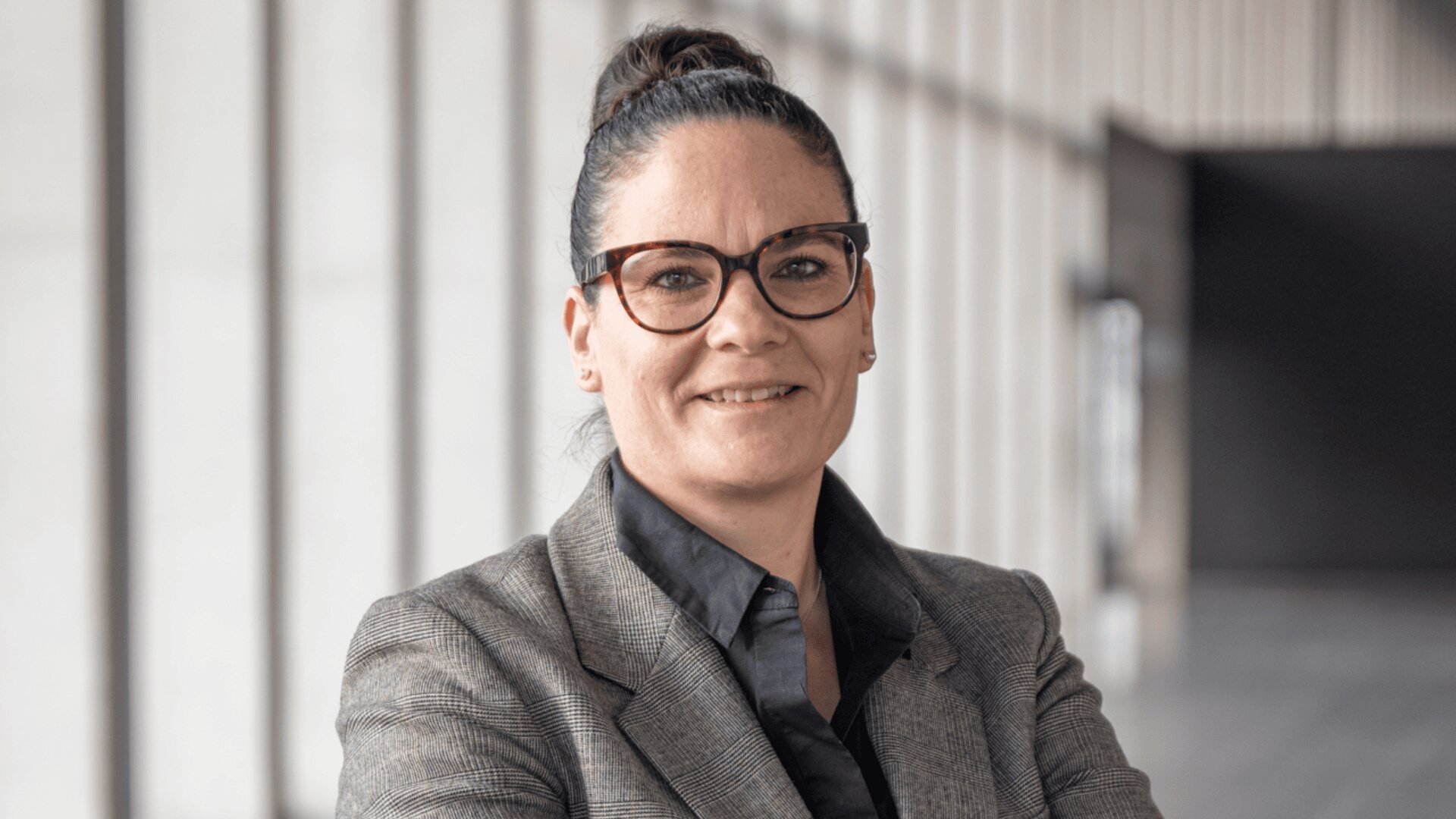 Eine Frau mit dunklem Haar in einem Dutt, einer Brille, einem grau-karierten Blazer und einem dunklen Hemd steht selbstbewusst mit verschränkten Armen in einem modernen, sonnendurchfluteten Flur mit hohen Fenstern.