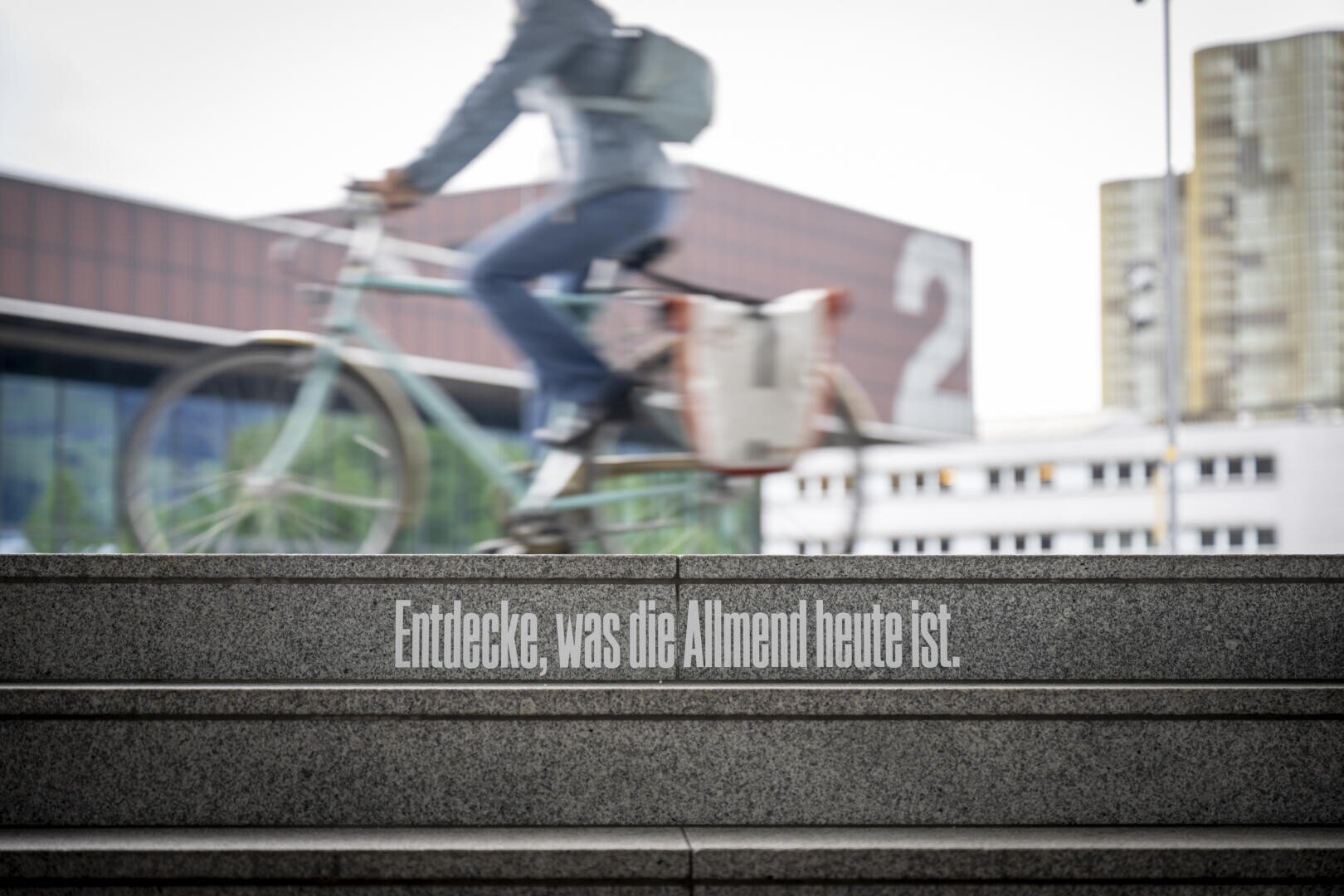 Eine Person fährt mit dem Fahrrad an verschwommenen Stadtgebäuden vorbei, die über Steinstufen zu sehen sind, auf denen der deutsche Text steht: Entdecke, was die Allmend heute ist.