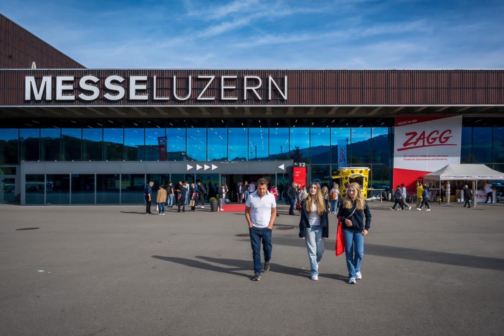Vor dem Messezentrum „Messe Luzern“ gehen Menschen vorbei, während sich am Eingang Gruppen versammelt haben und der Himmel klar ist. An dem Gebäude sind große Schriftzüge mit den Aufschriften „MESSE LUZERN“ und „ZAGG“ zu sehen.