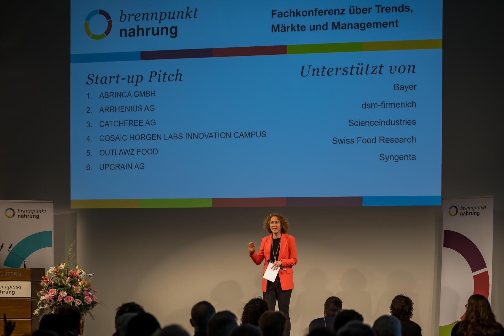 Eine Frau in einem roten Blazer spricht auf der Bühne einer Konferenz. Hinter ihr ist ein großer Bildschirm mit einer Liste von Startup-Pitches und Sponsoren zu sehen, vor ihr sitzt das Publikum.