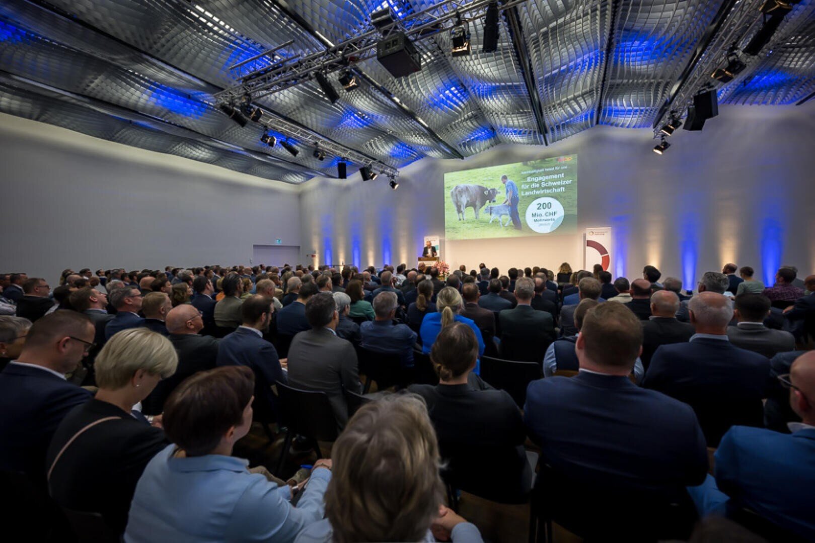 Ein großes Publikum sitzt in einem modernen Konferenzsaal und verfolgt, wie ein Redner bei blau-weißer Beleuchtung auf einer Leinwand Informationen über Kühe und Milch präsentiert.