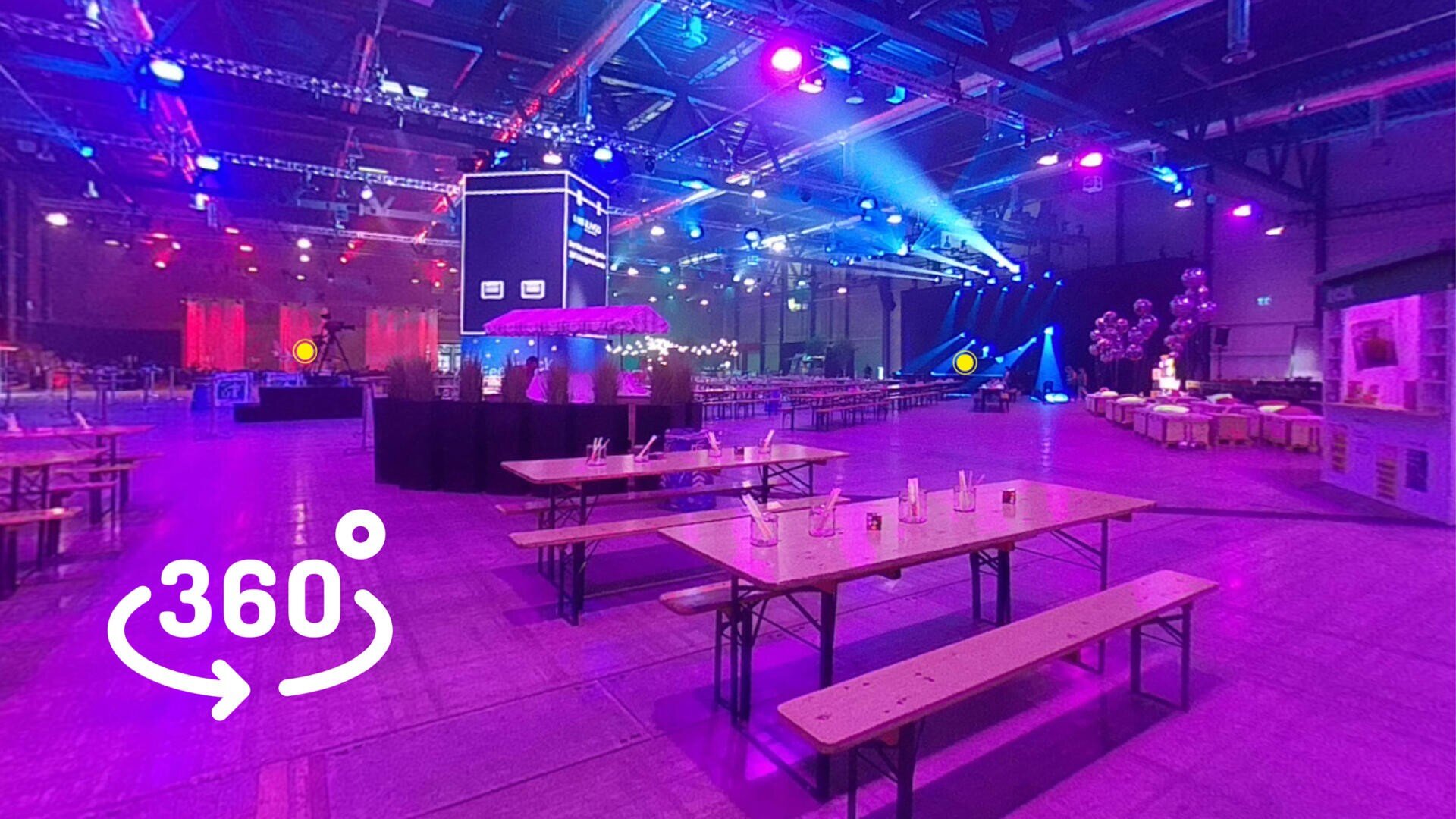 Ein großer Indoor-Veranstaltungsraum mit farbenfroher Bühnenbeleuchtung, Holztischen und -bänken, Luftballons und digitalen Bildschirmen. Im Vordergrund ist ein 360-Grad-Symbol sichtbar, das eine interaktive Panoramaansicht anzeigt.
