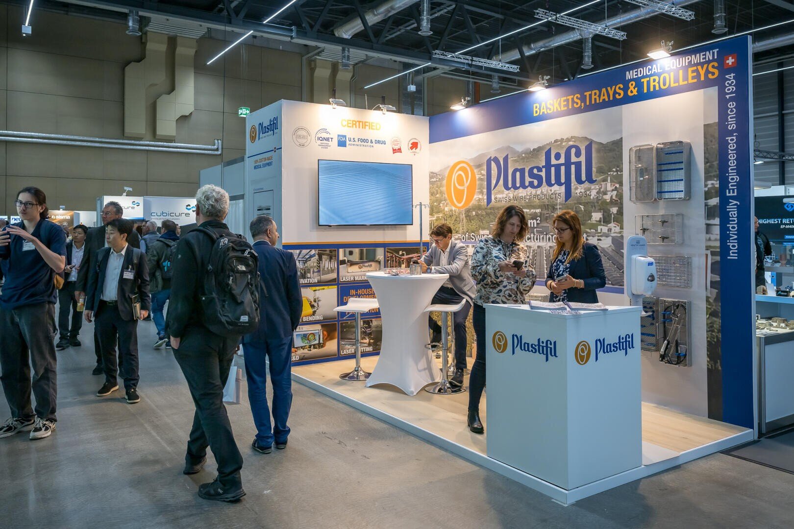 Besucher besuchen einen Messestand von Plastifil, einem Unternehmen, das medizinische Geräte wie Körbe, Tabletts und Wagen ausstellt. Zwei Frauen stehen hinter der Theke und unterhalten sich, während die Besucher vorbeigehen und sich die Ausstellung ansehen.