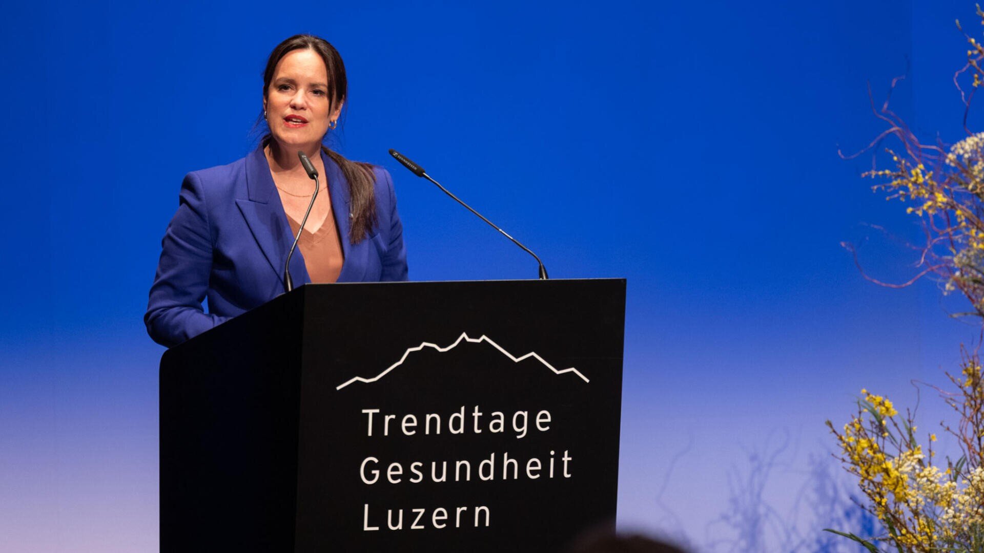 Eine Frau im blauen Blazer spricht an einem Podium mit dem Text „Trendtage Gesundheit Luzern“ auf blauem Grund, seitlich sind dekorative Pflanzen zu sehen.