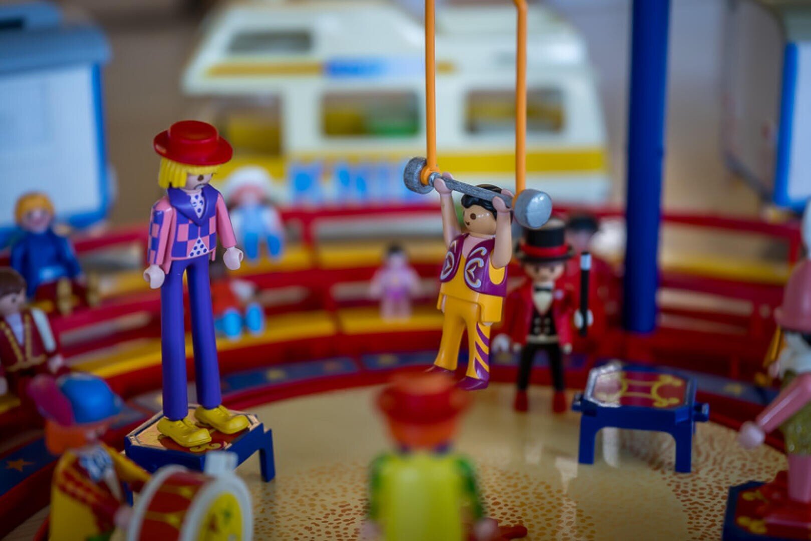Eine Nahaufnahme von bunten Spielzeugfiguren, die in einer Zirkusszene angeordnet sind, mit einem Trapezkünstler auf Schaukeln, Darstellern in bunten Kostümen und einem Clown auf Stelzen in einer Spielzeugzirkusmanege mit gelbem und blauem Hintergrund.