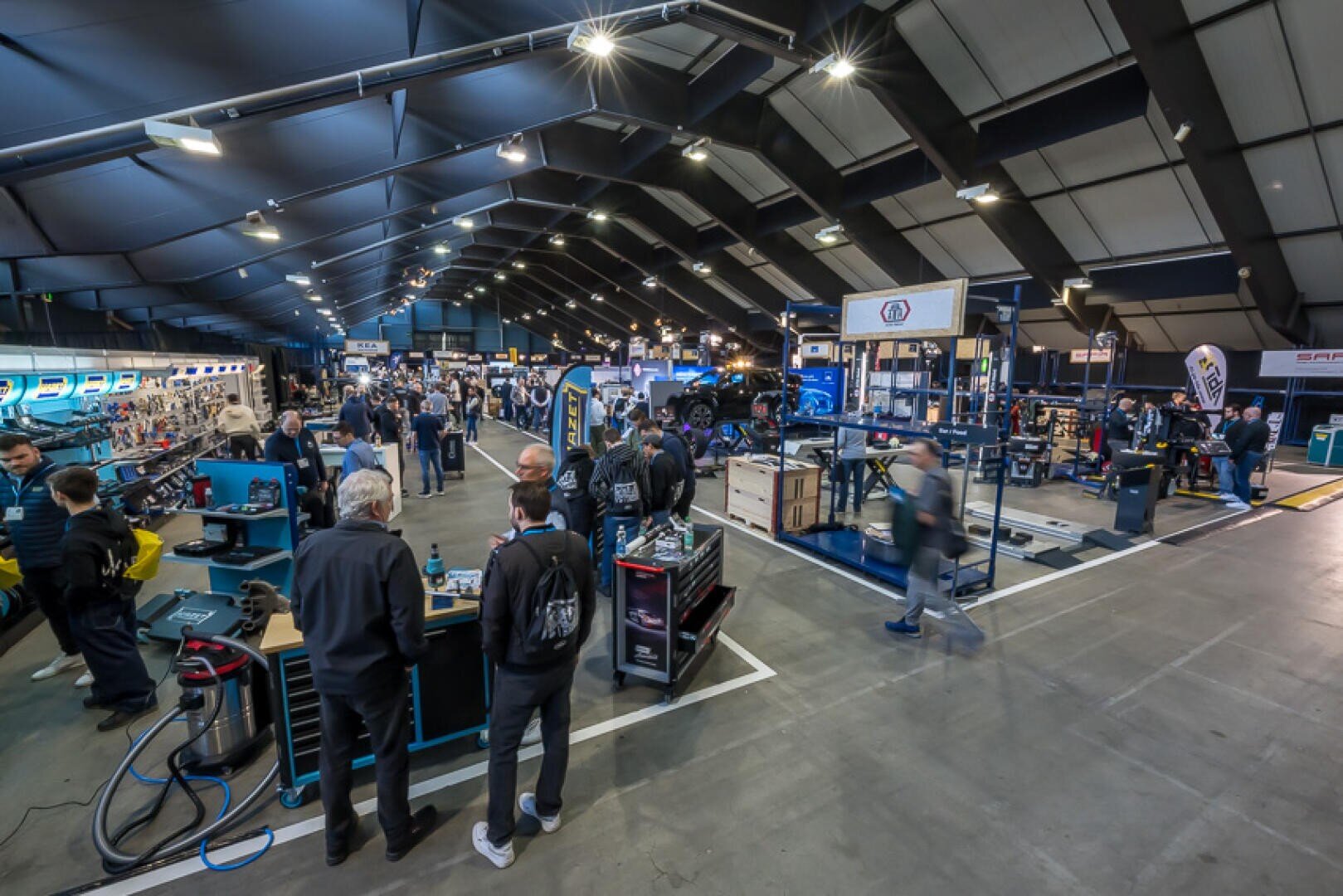 Eine große überdachte Ausstellungshalle mit Menschen, die umhergehen und an verschiedenen Ständen mit Werkzeugen, Ausrüstung und Fahrzeugen für die Automobilindustrie unter hellen Lichtern interagieren. Die Szene ist geschäftig und industriell, mit Werkstatteinrichtungen und Displays.