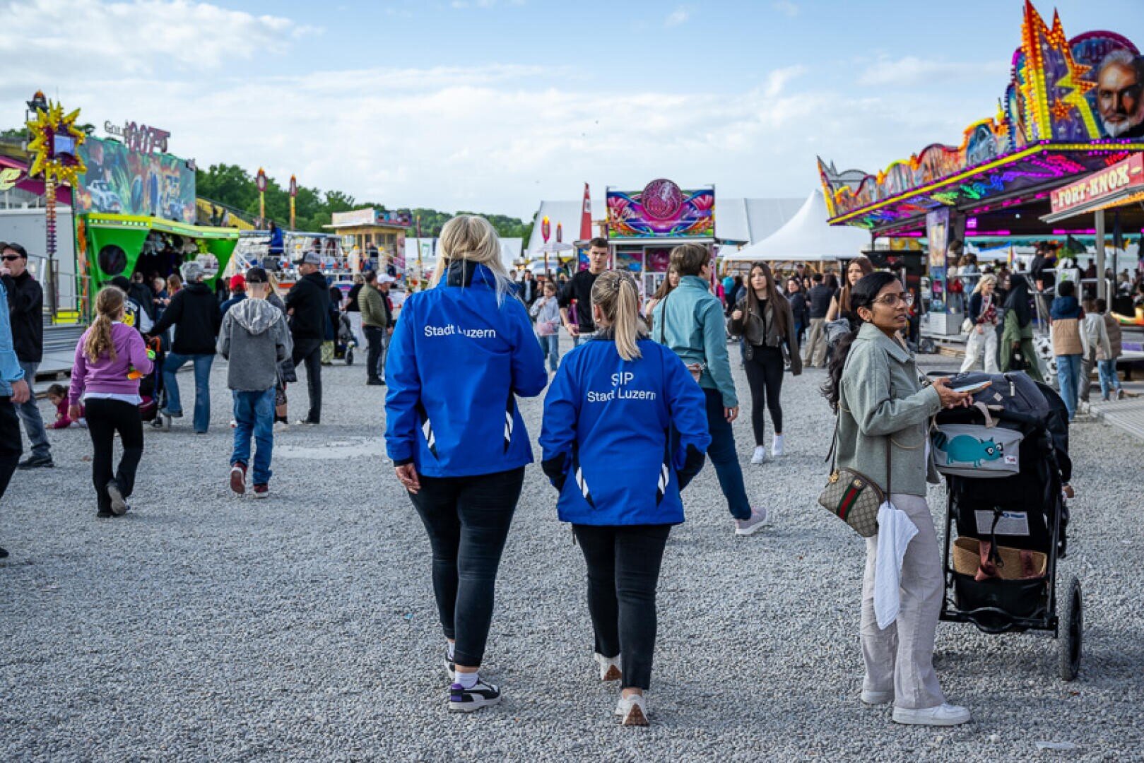 Zwei Personen in blauen Jacken mit der Aufschrift "Stadt Luzern" gehen über einen lebhaften Jahrmarkt mit Essensständen, bunten Fahrgeschäften und Menschenmassen. Eine Frau schiebt einen Kinderwagen auf dem Kiesweg unter einem hellen, teilweise bewölkten Himmel.