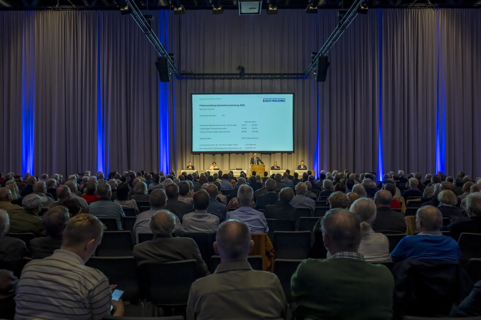 Ein großes Publikum sitzt vor einer Bühne mit mehreren Rednern und einer großen Projektionsfläche, auf der eine Präsentation in einem blau beleuchteten Konferenzsaal gezeigt wird.