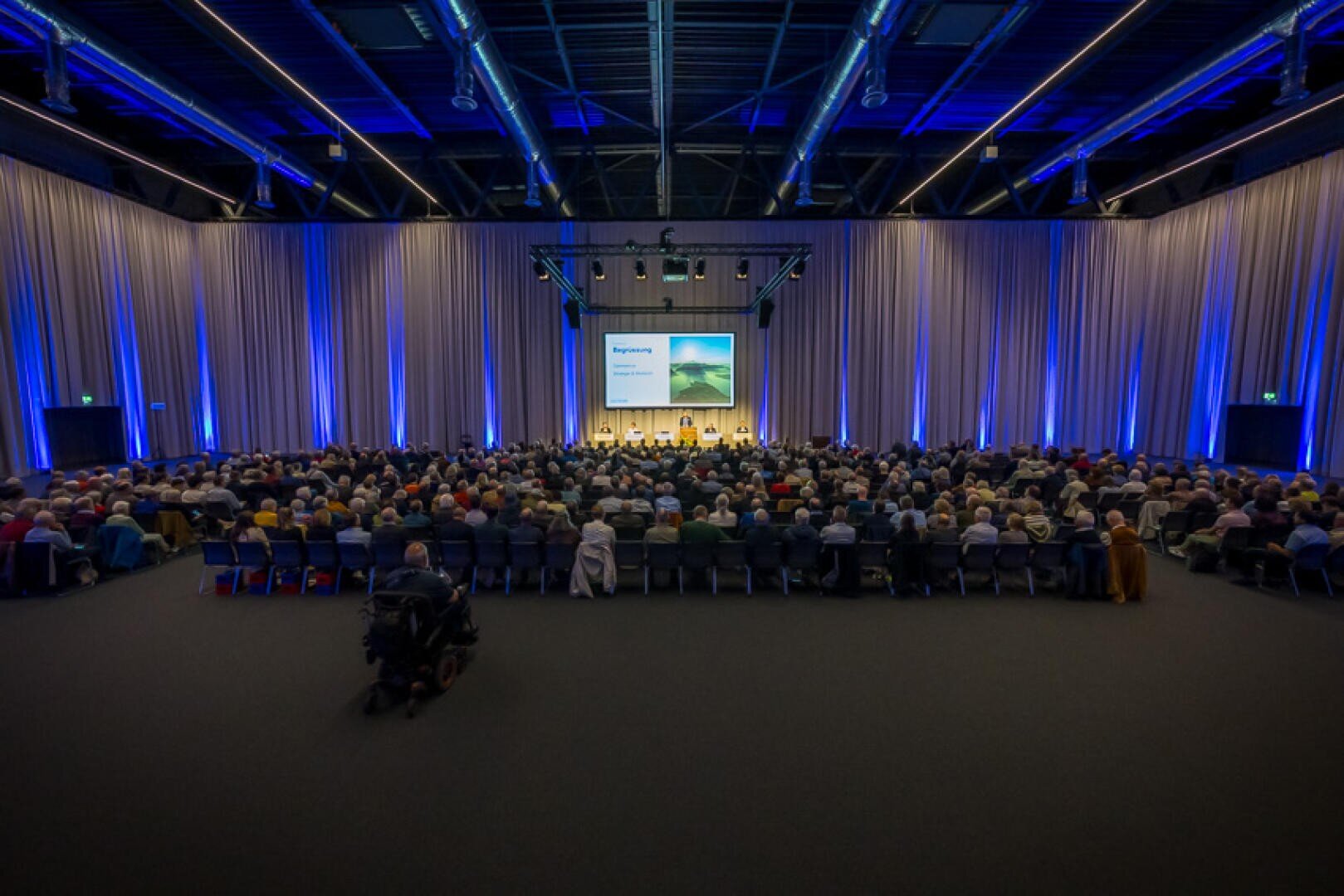Ein großes Publikum sitzt in Reihen vor einer Bühne mit Leinwand und Podiumsteilnehmern in einem geräumigen, modernen, mit blauem und weißem Licht beleuchteten Konferenzsaal.