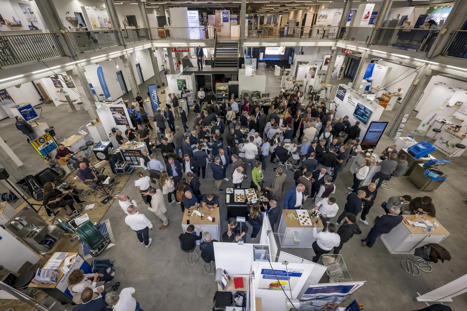 Aufnahme aus der Vogelperspektive einer belebten Messehalle mit verschiedenen Messeständen, Ausstellungsstücken und Menschen, die sich in einem mehrstöckigen Industriegebäude austauschen.