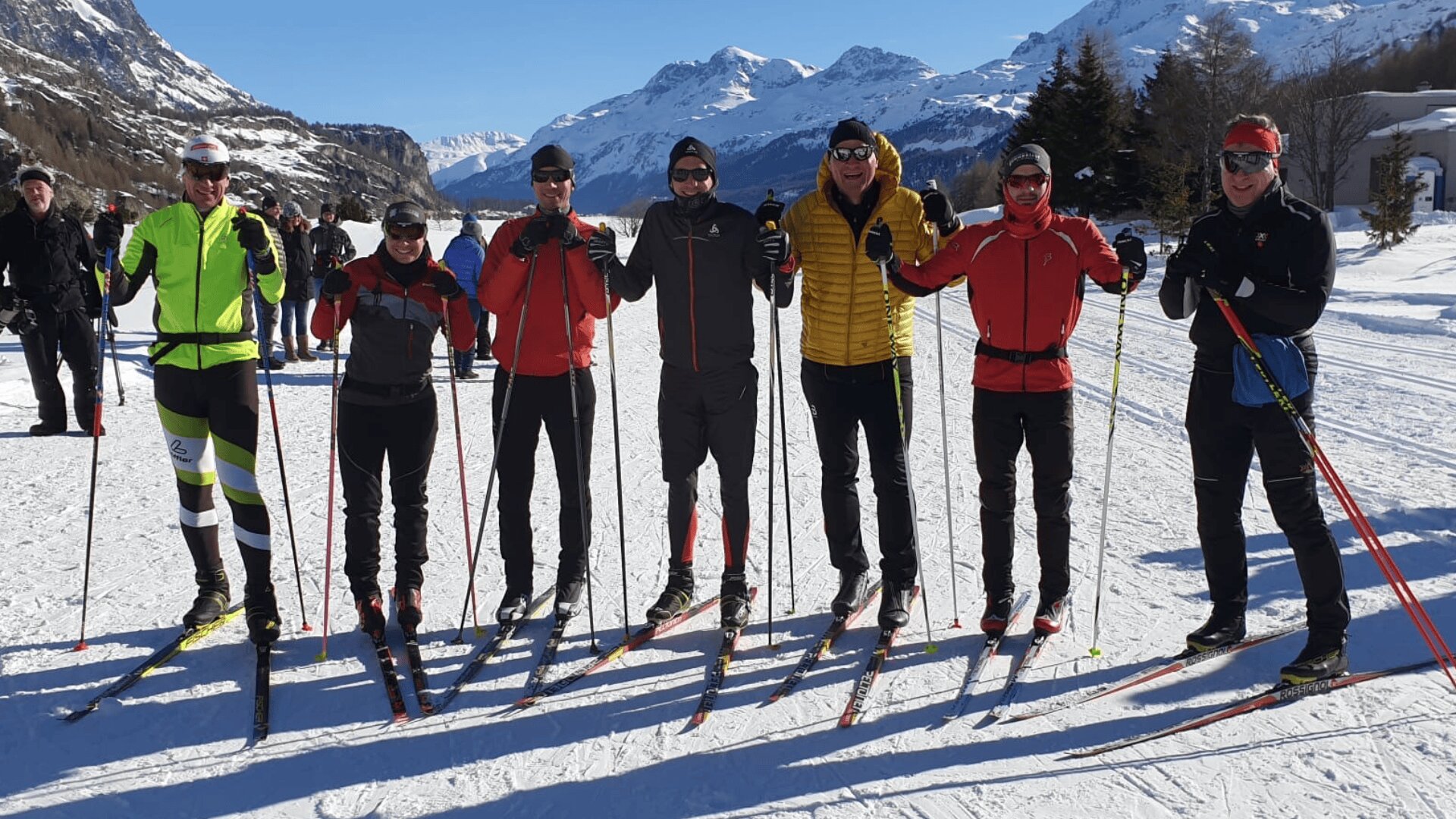 Sieben Menschen in Wintersportkleidung und Sonnenbrillen stehen mit Langlaufskiern Seite an Seite im Schnee, lächeln und haben die Arme umeinander gelegt, im Hintergrund sind schneebedeckte Berge und ein blauer Himmel zu sehen.