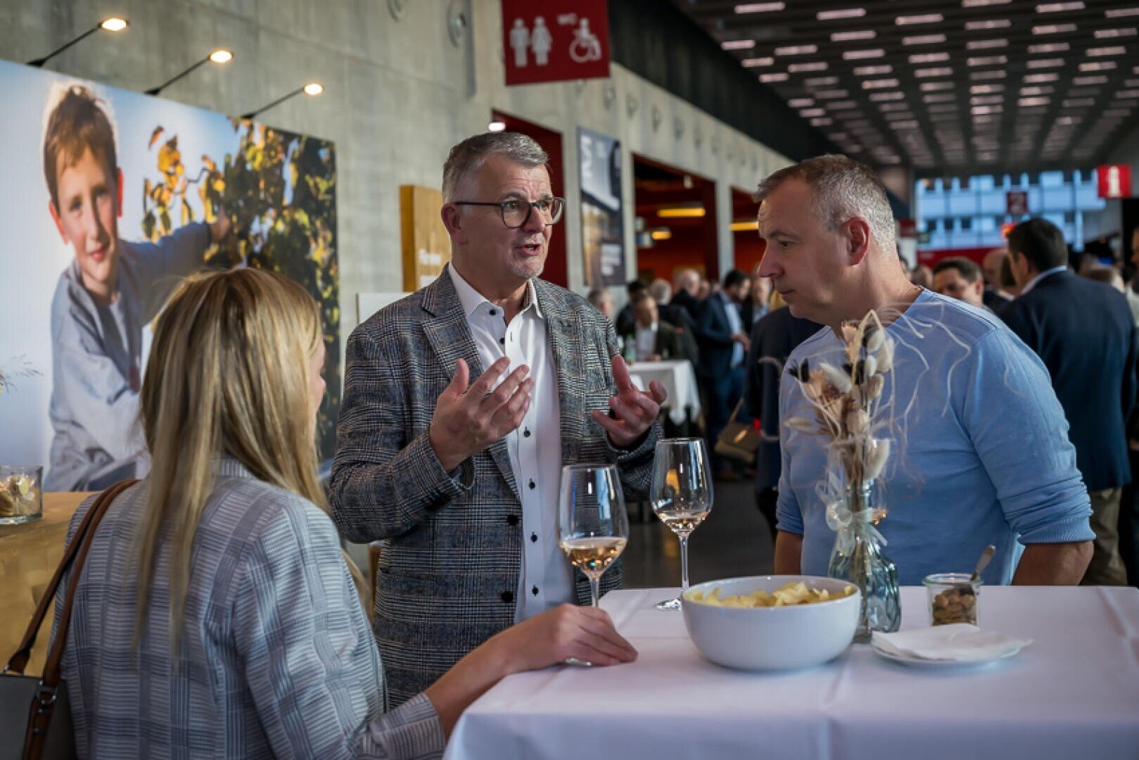 Drei Personen stehen um einen hohen Tisch mit Weingläsern und Snacks bei einer gesellschaftlichen Veranstaltung und unterhalten sich. Im Hintergrund sind andere Teilnehmer und ein großes Foto eines Kindes zu sehen.