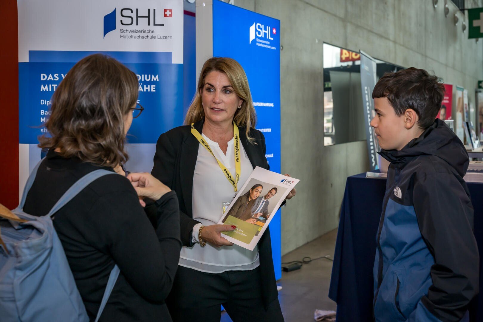 Eine Frau mit einer Broschüre spricht mit einer jungen Frau und einem Jungen an einem Messestand der SHL Schweizerische Hotelfachschule Luzern, mit Informationsbannern im Hintergrund.