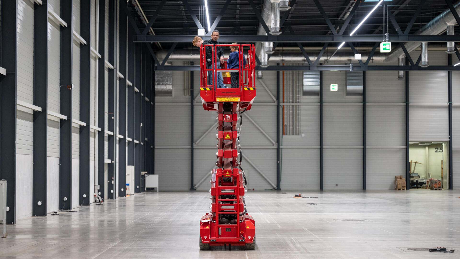 Drei Personen stehen auf einer roten Scherenhebebühne inmitten einer großen, leeren Industriehalle mit hohen Decken und grauen Wänden.