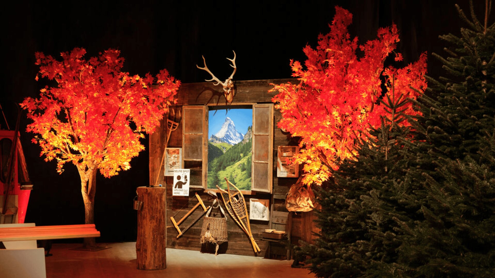 Eine rustikale Holzszene mit Hirschgeweihen über einem Fenster mit Blick auf die Berge, flankiert von leuchtend orangefarbenen Herbstbäumen und einer Kiefer, herumgehängt mit Holzwerkzeugen und Postern.