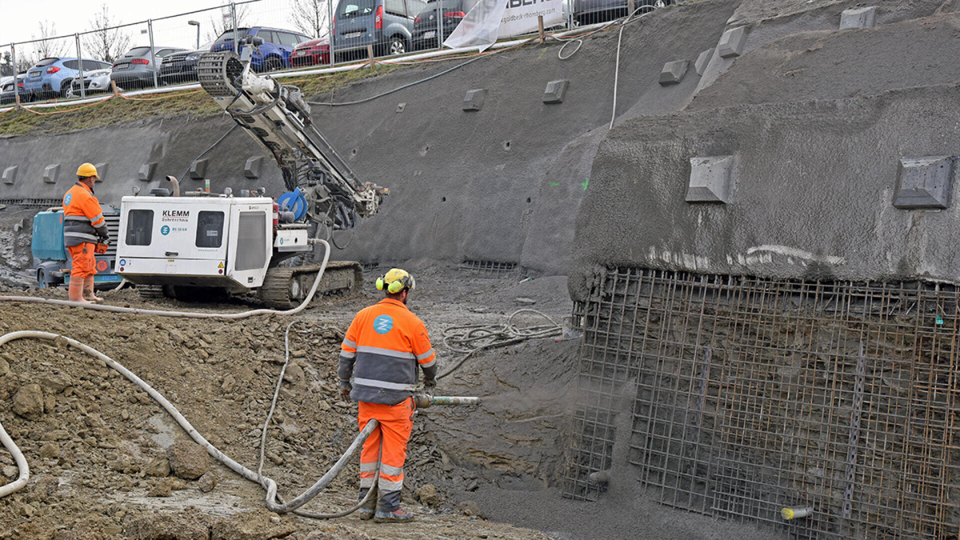 Zwei Bauarbeiter in orangefarbener Schutzkleidung spritzen Beton auf eine in einen Erdhang eingelassene Drahtgeflechtkonstruktion, während in der Nähe Maschinen am befestigten Hang arbeiten.