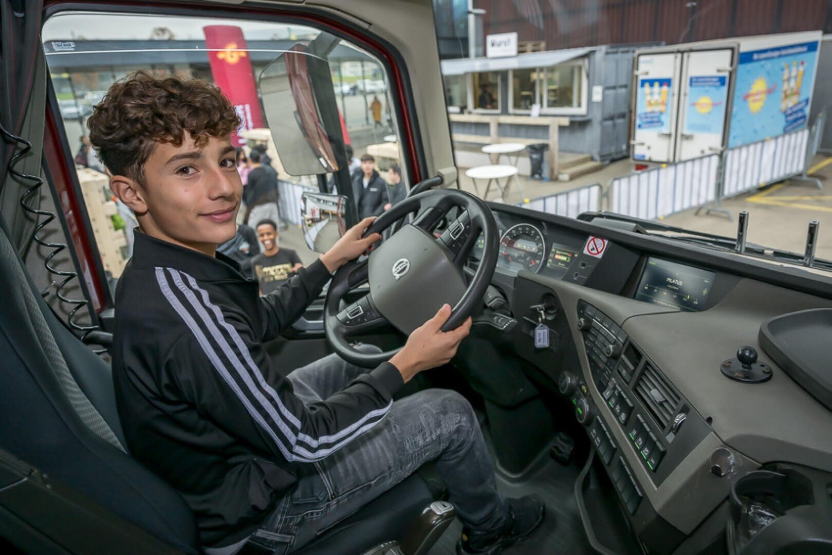 Ein lächelnder Jugendlicher sitzt auf dem Fahrersitz eines großen Lastwagens und hält das Lenkrad fest. Die Tür des Lastwagens ist offen, und draußen sind Menschen in der Nähe von Gebäuden und Informationsdisplays zu sehen.