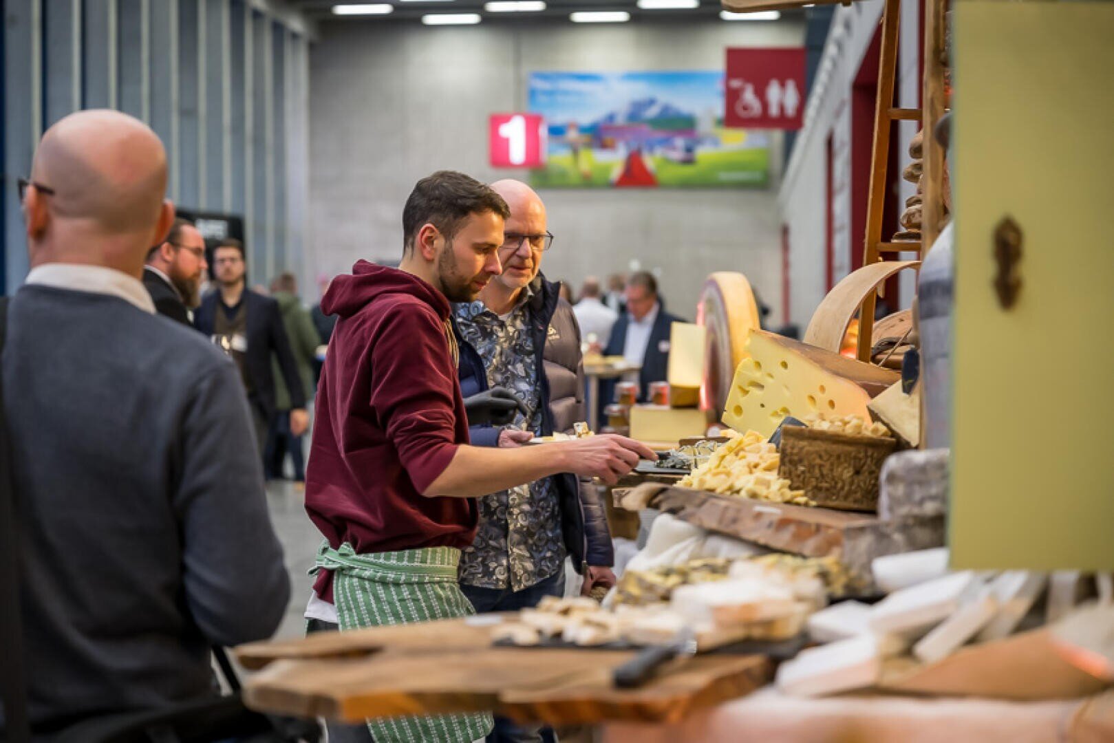 Mehrere Personen stehen an einer Käsetheke in einer Markthalle, probieren und wählen Käse aus. Es herrscht reges Treiben, denn die Käselaibe und geschnittenen Stücke sind auf Holzflächen ausgestellt.