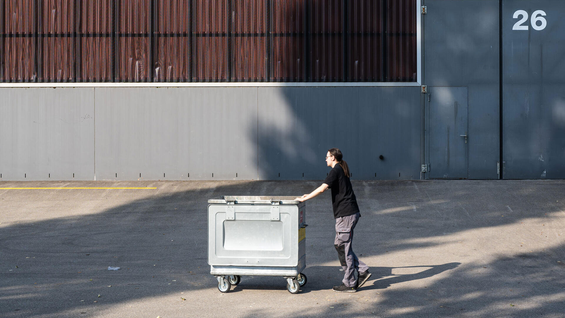 Eine Person in einem schwarzen Hemd schiebt eine große graue Mülltonne auf Rädern über einen leeren, gepflasterten Platz vor einem grauen Industriegebäude mit der Nummer 26.